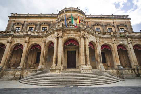 View of the historic Palazzo Ducezio with its elegant Baroque facade and arched portico under a cloudy sky in Noto, Sicily, Italy.