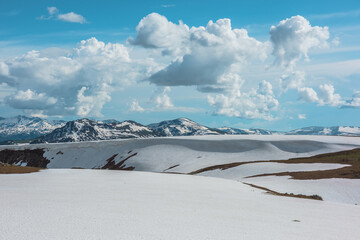 Scenic landscape with sunlit snowfield against snow cornice and big snowbound mountain range far away under dramatic cloudy sky. Vast view with snowy glade in high mountains under clouds in blue sky. © Daniil