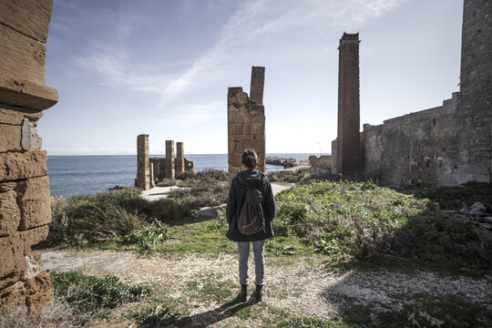 View of a woman standing among the historic stone ruins and tall chimney of the Tonnara di Avola overlooking the Mediterranean Sea in Avola, Sicily, Italy.
