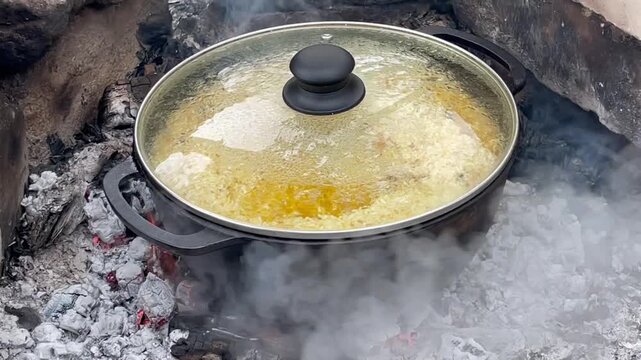 Traditional pilaf cooking in a large cast-iron cauldron over an open fire supported by bricks outdoors.