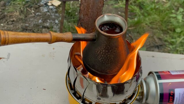 Brewing coffee in a copper cezve over an open flame on a portable gas stove outdoors.