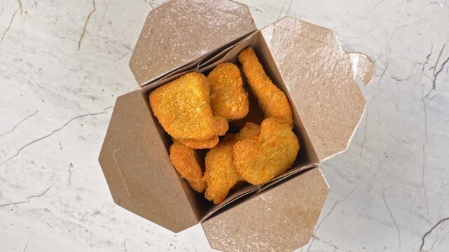 Hand grabbing a crispy brown chicken nugget from an open brown paper takeout delivery box top down view
