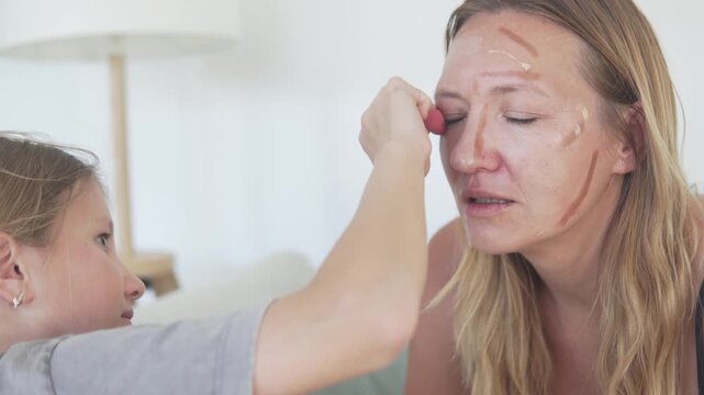 Little daughter playfully applying makeup to her mother with a cosmetic sponge at home. Authentic family beauty routine with contour lines, natural emotions, self-care, humor and warm mother-daughter