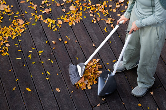 A woman is cleaning fallen autumn leaves on a wooden terrace using a plastic broom and dustpan. Top view.
