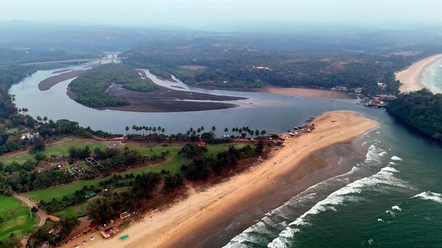 Cinematic aerial view of a luxury beachfront resort surrounded by expansive greenery, calm backwaters, and the Arabian Sea, with waves tracing the shoreline beneath a soft hazy sunset atmosphere.