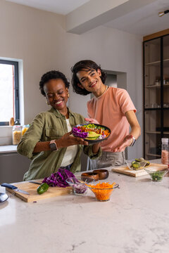 Diverse friends smiling and offering salad bowl in home kitchen with purple cabbage avocado carrots