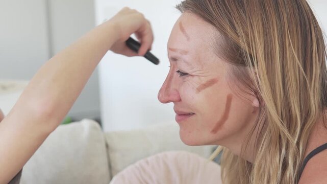 Little daughter applying makeup to her smiling mother at home with a cosmetic brush. Authentic family beauty routine showing playful self-care, natural skin texture, contour lines and warm mother