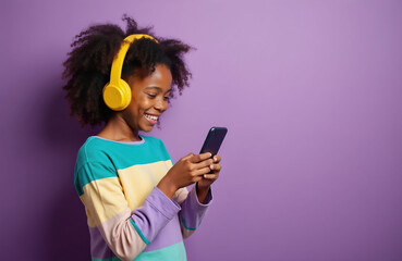 Naklejka premium Young Black girl wears yellow headphones, laughs while playing game on phone. She has curly hair and a colorful shirt. Isolated on purple background.