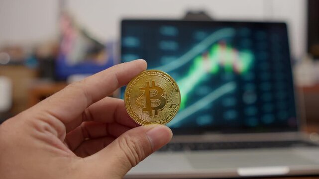Close-up of a hand holding a gold Bitcoin coin in front of a cryptocurrency market chart, illustrating digital investment, trading, and financial volatility.