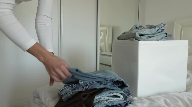 Close-Up of Female Hands Folding Light Blue Denim Jeans Above Pile of Mixed Pants Next to White Storage Box on Bed &mdash; Woman Sorting Casual Clothing in Bright Minimalist Bedroom, Wardrobe Declutter
