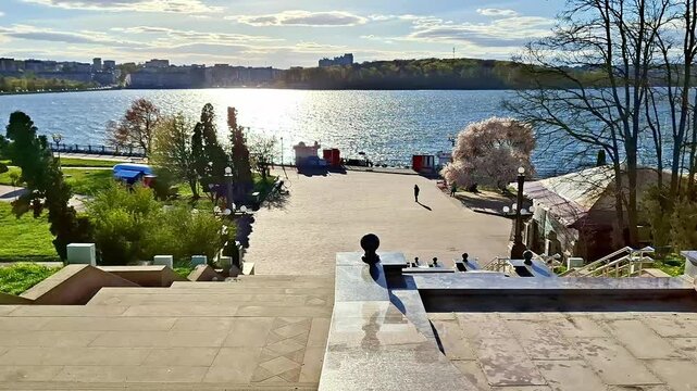 Stone steps lead to a tree lined promenade beside sunlit water, with kiosks and distant shoreline buildings creating a calm and picturesque urban waterfront view
