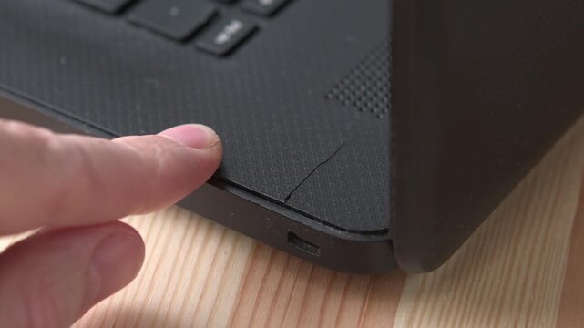 Finger shows cracked laptop casing as stiff hinge strains during repeated opening and closing on wooden desk in closeup view