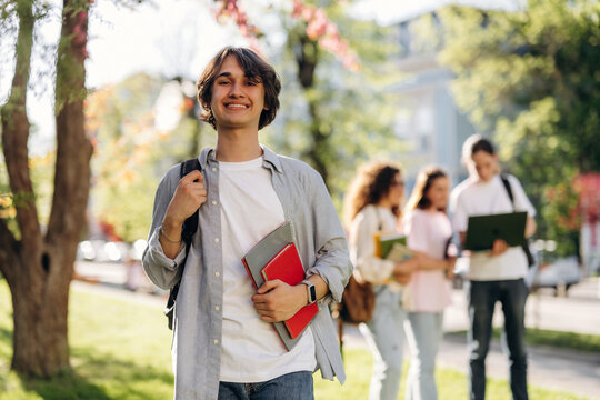 Young male student with backpack smiles while holding books in a sunny park, with classmates discussing in the background near trees and buildings