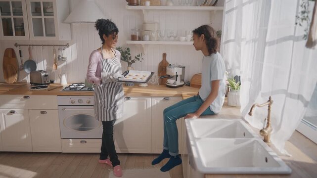 Cheerful black woman holding hot baking sheet with cookies, talking with girl. Mother and daughter cooking sweets at home, enjoy delicious hypoallergenic homemade bakery, traditional healthy food