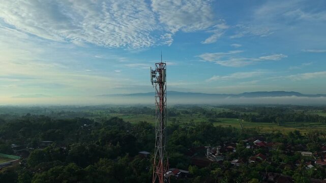 Aerial view of a telecommunications tower overlooking a rural village and misty mountains under a blue sky in Kemalang, Central Java, Indonesia.