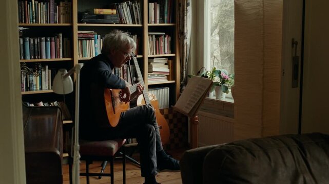 Senior musician practicing acoustic guitar in his cozy living room with a large bookcase