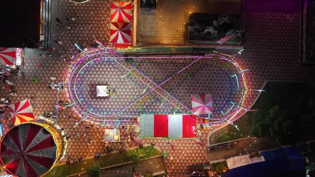 Aerial view of GOR Gelarsena Klaten Night Market with a neon-lit infinity track ride, striped tents, and people at night, Kecamatan Klaten Utara, Jawa Tengah, Indonesia.