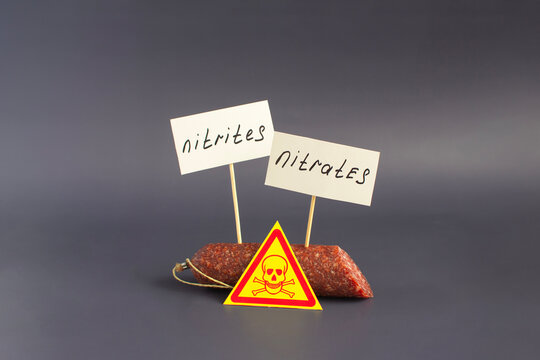 A piece of smoked sausage, a danger sign and warning labels "Nitrates and nitrites" in close-up on a dark background