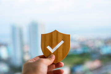 Hand holding wooden shield with checkmark against blurred city background. Concept of safety, security, quality approval, trust, and real estate insurance. Ideal for business protection. © sommart