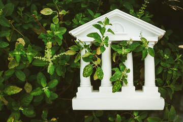 Naklejka premium White bank building model overgrown with green plants. Outdated financial system, abandoned institution, economic crisis, nature reclaiming architecture, and obsolete business concept.
