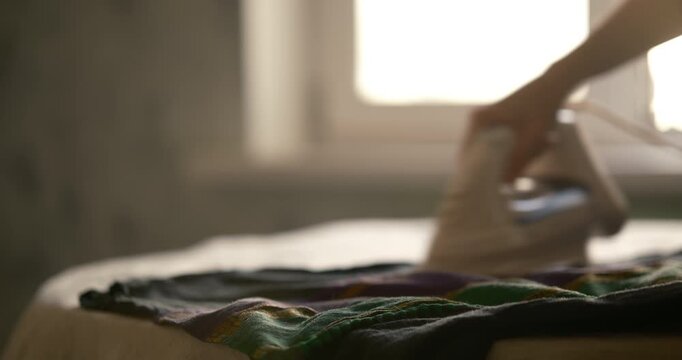 Cinematic close up iron on table and a black sweater with an Indian pattern. Blurred hands ironing the sweater in the background. Cozy home atmosphere and household routine. Window and home interior 