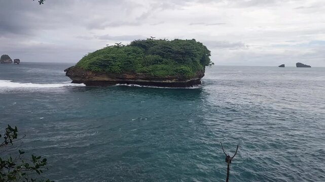 Small tropical island with lush greenery surrounded by turquoise ocean under cloudy sky, located at Tanjung Penyu beach, East Java, Indonesia.