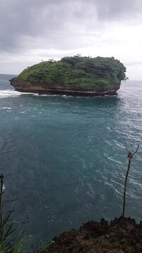 Small tropical island with lush greenery surrounded by turquoise ocean under cloudy sky, located at Tanjung Penyu beach, East Java, Indonesia.