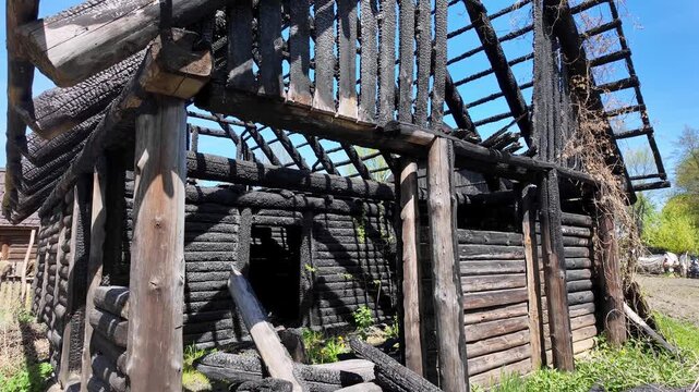 Burnt wooden log house, charred roof structure and black walls after a fire, visible burnt wood textures, ruins against a blue sky and green grass in bright sunlight, building destruction details.