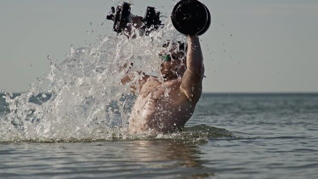 Dumbbells training creates beach workout. Muscular man uses dumbbells outdoors. Dumbbells exercise builds shoulder strength. Athlete trains with dumbbells near ocean.