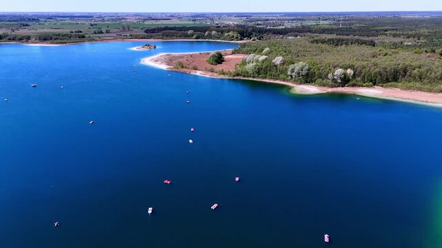 Aerial drone view of Niedzięgiel Lake in Skorzęcin, vibrant blue water with numerous pedal boats and small crafts, lush green forests lining sandy shores under a clear sunny sky with horizon panorama.