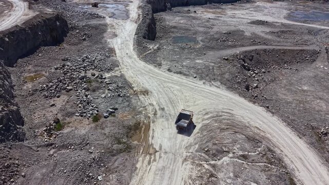 A truck drives on a dirt road at a mining site. The area is rocky with large mounds of dirt and few water pools. The landscape shows evidence of active quarry operations in progress.