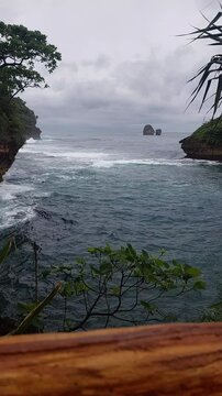 Beautiful ocean inlet surrounded by tropical cliffs and lush greenery with distant rocky islands under cloudy skies at Tanjung Penyu beach, East Java, Indonesia.