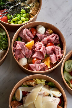 Variety of gourmet salad bowls in craft paper packaging on a white marble surface. Assortment of healthy takeout meals including roast beef, salmon, and chicken.