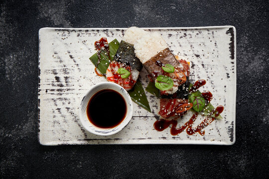 Gourmet spam musubi served with soy sauce and chili on a rustic ceramic plate. High-end Asian fusion photography on a dark stone background.