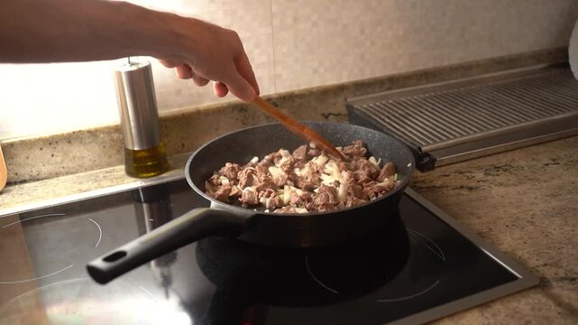 Male hand stirring diced meat and onion with a wooden spoon in a pan on an induction stove