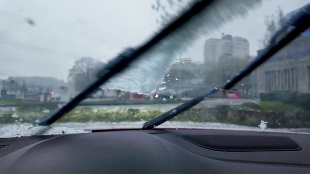 Gros plan sur des petits gr&ecirc;lons qui tombent sur le parebrise depuis l'int&eacute;rieur d'une voiture. Les essuie glaces les balayent. Boules de glace apr&egrave;s un orage de printemps.