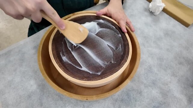 Mains d'une femme faisant passer une pr&eacute;paration de colle avec une spatule en bois dans un tamis afin d'&eacute;liminer les grumeaux. Geste artisanal, r&eacute;p&eacute;titif et m&eacute;ticuleux.