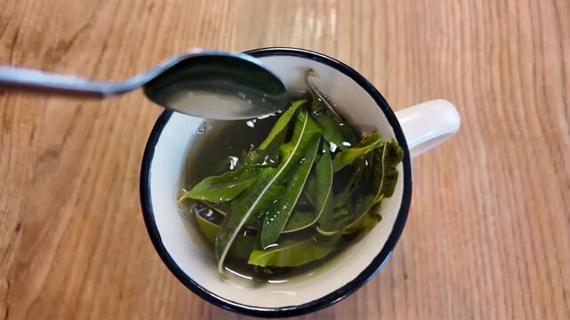 Une tisane aromatique avec des feuilles de verveine citronn&eacute;e d'o&ugrave; s'&eacute;chappe une vapeur chaude, pos&eacute;e sur une surface en bois avec une main qui m&eacute;lange les feuilles avec une cuill&egrave;re.