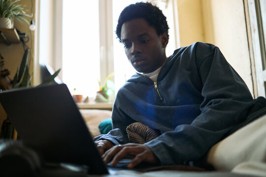 African American teenage boy using laptop while lying on sofa at home, studying online and browsing digital content. Useful for remote learning, youth technology, screen time campaigns