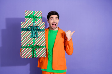 Naklejka premium Excited young man in orange jacket holding stacked Christmas gifts and pointing with joy at colorful holiday presents