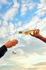 Obraz premium Hand pouring white wine into glass with cloudy blue sky background. Concept of vineyard tourism, wine tasting, romantic getaway, premium beverage branding and outdoor dining.