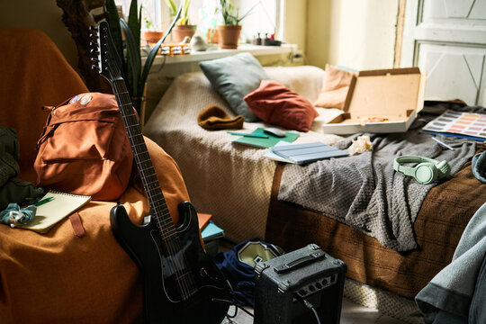 Wide shot of teenager bedroom showing guitar, amplifier, headphones, notebooks, backpack, and pizza box, suggesting creative student lifestyle. Useful for youth culture or study life marketing