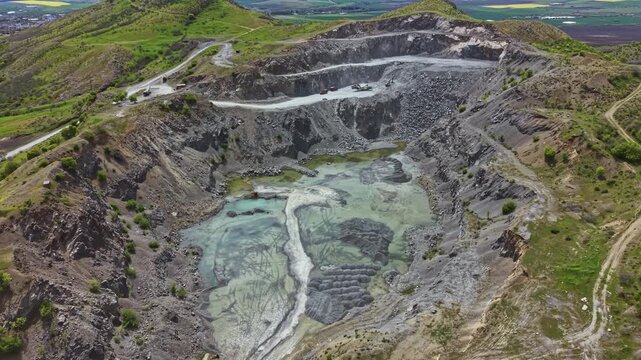 A large open pit mining site shows a deep excavation with water collected at the bottom. Surrounding hills frame the area while machinery is visible working on the site.