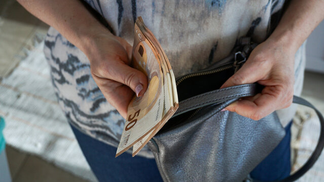 Woman placing euro banknotes into small handbag representing personal finance, saving habits, budgeting, cash handling and everyday financial management in domestic context
