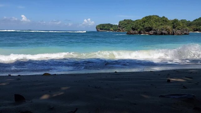 Turquoise ocean waves crashing on sandy beach with small tropical island view, located at Gatra beach, East Java, Indonesia.