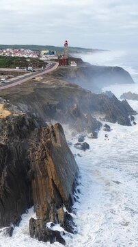 Aerial view of Farol Penedo da Saudade standing on rugged cliffs with crashing waves and a coastal road in Marinha Grande, Leiria, Portugal.