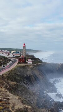 Aerial view of Farol Penedo da Saudade lighthouse standing on rugged cliffs overlooking the Atlantic Ocean and coastal road in Marinha Grande, Leiria, Portugal.