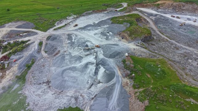 Heavy machinery works on a quarry site with piles of gray material spread across the ground. Green hills surround the area under bright daylight with open fields nearby.