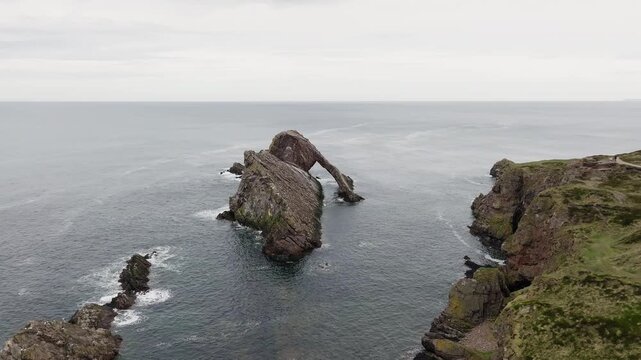 Drone footage flying toward Bow Fiddle Rock, Portknockie, Moray coast, Scotland, overcast day, dramatic sea arch, dark choppy waters, moody grey sky above