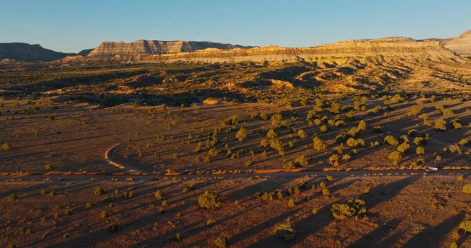 Truck camper driving on winding dirt road curves across desert plain with mesas near Escalante Utah during golden hour, drone static